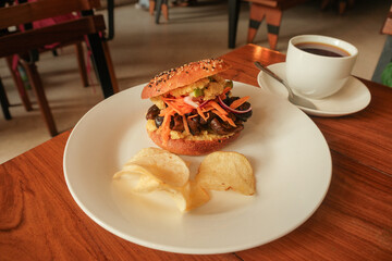 Travel photography in Asunción, Paraguay. High angle close up shot of gourmet vegetarian sandwich bagel with mushrooms; chips served on white plate in cozy café setting. Coffee on table in background.