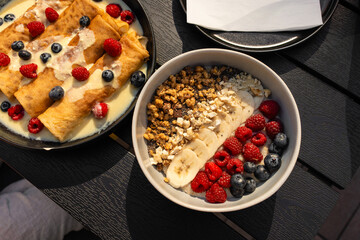 Flat lay of european breakfast served outdoors, featuring sweet pancakes with vanilla sauce and berries alongside a healthy bowl with banana, granola, raspberries and blueberries.