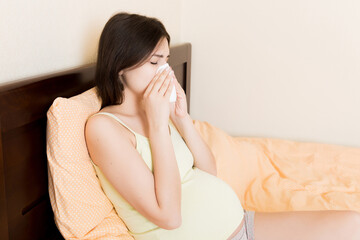 Portrait of young unhappy pregnant woman leaning on pillows on bed and blowing her nose into...