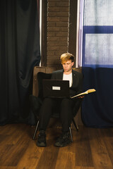 Focused young man sits in a chair, working on a laptop and reading a book. He is dressed in a...