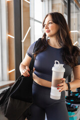 Curvy brunette woman in gym outfit holding sports bag and protein drink at modern fitness studio