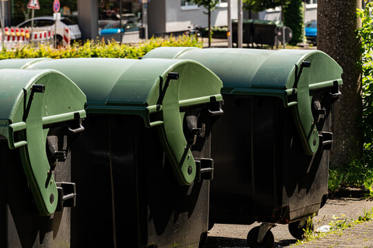 A group of green trash bins is lined up on the sidewalk of an urban area. The sun shines brightly, illuminating the bins and surrounding greenery. City buildings are visible in the background.