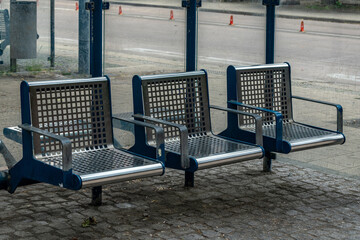 Three modern metal benches are positioned at a bus stop in an urban area. The benches feature a...