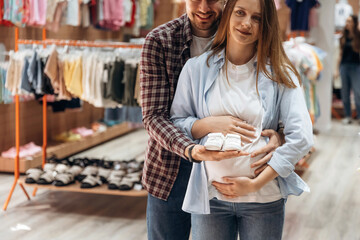 White cute little shoes. Pregnant woman with man are in the kid's clothing store
