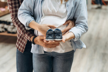 Embracing, holding little shoes. Pregnant woman with man are in the kid's clothing store