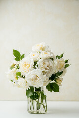 White rose bouquet in glass vase on white table with beige textured wall background