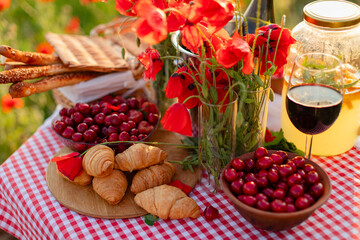 Summer picnic with croissants, cherries, lemonade, wine, and poppies on a red checkered tablecloth in a blooming poppy field at golden hour.
