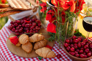 Close-up of fresh croissants, cherries, grissini, and poppy flowers on a picnic table with a red gingham tablecloth in a summer meadow.