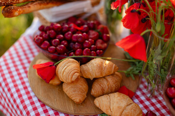 Close-up of fresh croissants, cherries, grissini, and poppy flowers on a picnic table with a red...