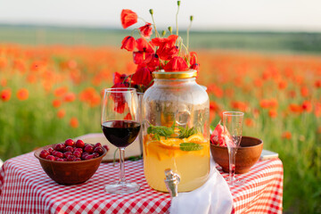 Close-up of summer lemonade with orange slices and mint in a glass jar, next to a glass of red wine and a bowl of cherries on a picnic table in a blooming poppy field.