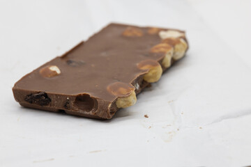 A piece of milk chocolate with hazelnuts and raisins on a white surface, highlighting texture and...