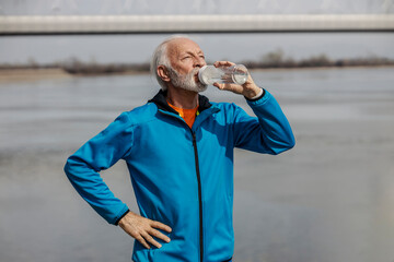 Portrait of thirsty senior sportsman drinking water near riverbank.