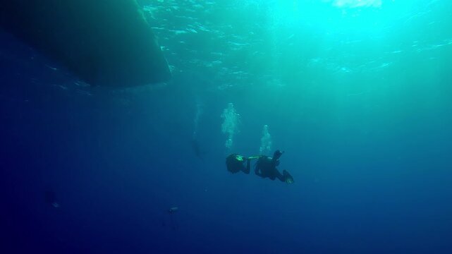 Scuba divers swim in the blue depths towards a diving boat with a sunny day in backlight, Slow motion of divers returning underwater to a safari boat