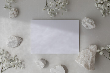 Neutral background with white decorated stones, blank paper card and gypsophila flowers in flat lay style. Spa or wedding invitation in minimalistic modern style.