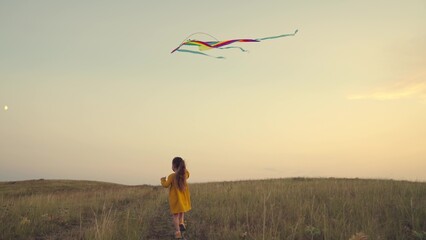 a little girl runs along the road catching wind in a kite at sunset in sky, childhood dream of...