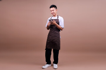 Full Length Of Smiling Young Asian Guy In Brown Apron Using Smartphone While Standing