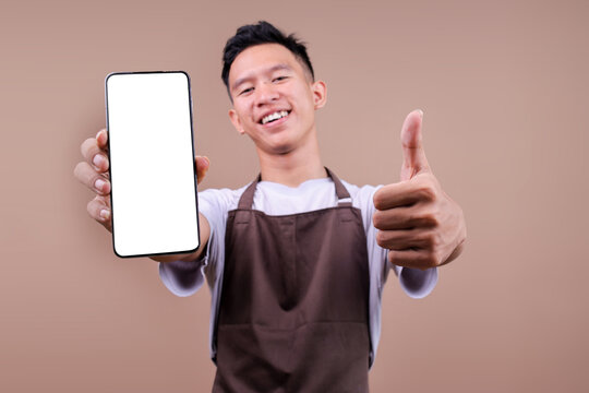 Smiling Young Man In Brown Apron Showing Blank Smartphone And Giving Thumbs Up Over Beige Background