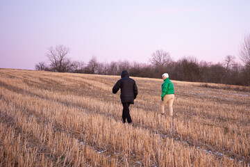 Couple enjoys a winter walk through a quiet field at dusk, surrounded by bare trees and frosty grass under a pastel sky
