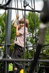 Obraz premium A child boy is climbing up an alpine grid in a park on a playground on a hot summer day. Kids playground in a public park, entertainment and recreation for children, mountaineering training.