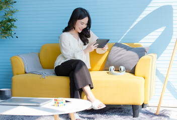 Asian young beautiful happy woman wears casual clothes smile holding using digital tablet technology computer sitting on yellow sofa in blue living room at home using apps shopping online reading news