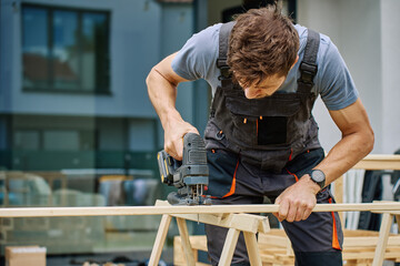 Man cutting wooden plank with electric jigsaw at home backyard. Outdoor carpentry and DIY work