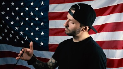 Man with full sleeve tattoo and black clothing lifting up a holy black cross in his arm on a chain, watches it and prays religiously while he is in front of an American flag. Cross moves around.