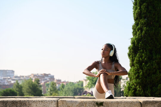 Young sporty woman stretching legs listening music with wireless headphones in city park
