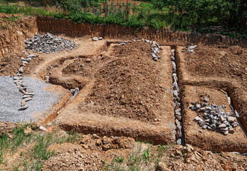 An outdoor construction site shows freshly dug trenches filled with rocks and gravel, marking the...