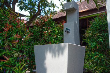 An outdoor sink with a metal faucet surrounded by green and red ornamental plants. The modern stone structure is situated in a shaded garden area, evoking cleanliness and nature.