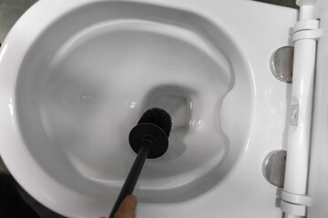 Hand holding a black toilet brush, scrubbing a white ceramic toilet bowl, promoting cleanliness and hygiene while tackling dirt and grime in the bathroom environment