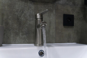 Modern bathroom faucet pouring water into sink with a gray tiled wall and black electric outlet in the background, promoting water conservation and modern bathroom design
