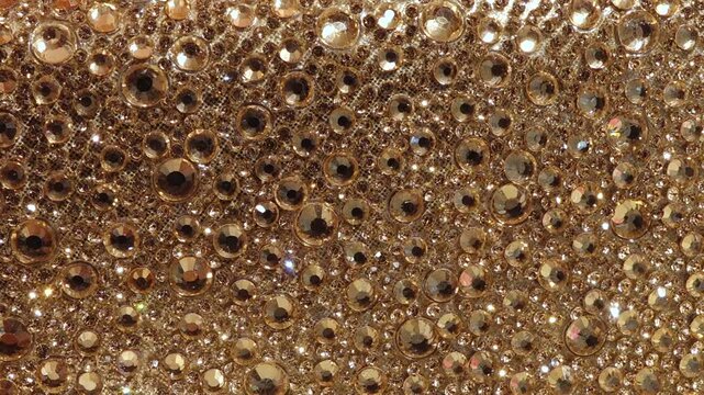 Macro shot of a decorative surface densely covered in shiny, gold rhinestones of various sizes, creating a rich, sparkling texture.
