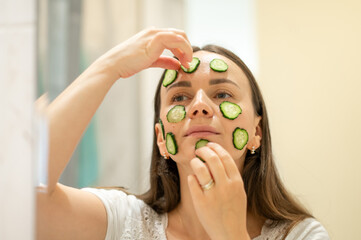 reflection of a woman in a mirror who puts cucumber slices on her face, face mask