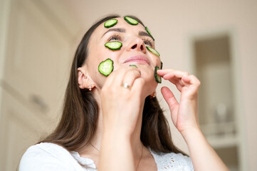 Woman with cucumber slices on her face enjoying a healthy skincare routine in a bright kitchen