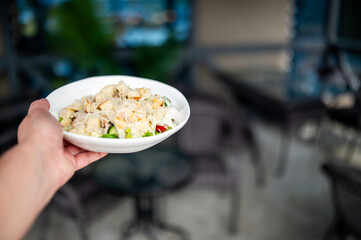 Hand holding a white bowl with Caesar salad, croutons, grated cheese, and lettuce. Blurred indoor background with tables and chairs. Fresh and appetizing presentation.