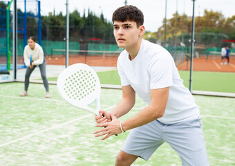 Portrait of sporty young guy hitting two handed backhand during paddle tennis friendly match in...