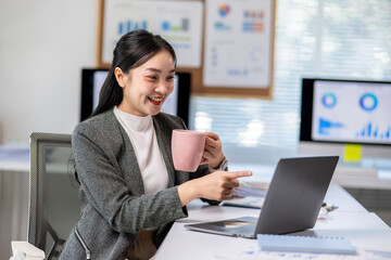 Fototapeta premium Asian businesswoman drinking coffee and pointing at laptop screen