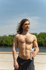 Shirtless fit brunette man with curly long hair holding a water bottle while standing outdoors on a...