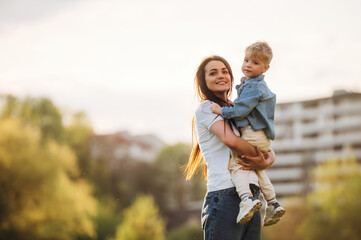 Cheerful, having fun, holding boy in hands. Mother and son are together on the summer field