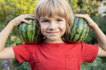 Portrait of a smiling boy's face 8 years old with two small striped watermelons on his shoulders....