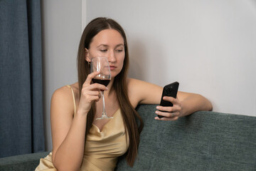 Young woman in elegant dress holding glass of red wine while using smartphone, sitting on...