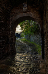 Obraz premium Evening light reflects on the cobblestones of a damp and mossy medieval path through an ancient stone archway with a religious statue at dusk with the last rays of the sun in Conques, France.