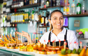 Cheerful Latin female bartender in uniform serving delicious tapas on skewers at the counter of the...