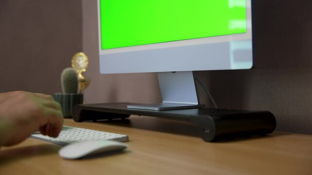 A person types on a wireless keyboard in front of a desktop monitor with a green screen. The camera slowly pans across a modern desk setup with a cactus.