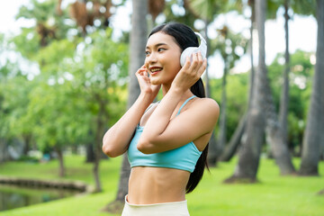 Asian sports woman listening to music on headphones outdoors. Female in sportswear standing against in park. Happy lady athlete taking a break from her workout