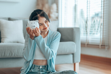 Young Asian woman holding piggy bank and sitting on the floor at home. Save money and financial investment