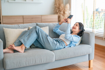 Happy cheerful positive young Asian woman on the phone at home, smiling girl using app and browsing on the internet on smartphone lying on sofa
