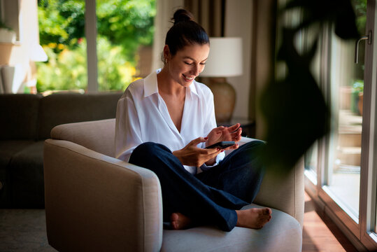 Smiling brunette haired woman relaxing in an armchair in her luxury home and using phone