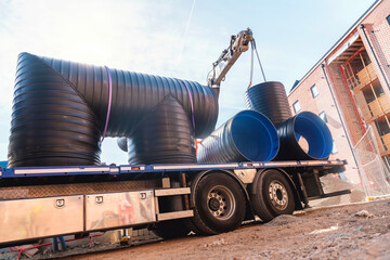 Construction workers maneuver a crane to lift oversized black plastic pipes from a truck at an...