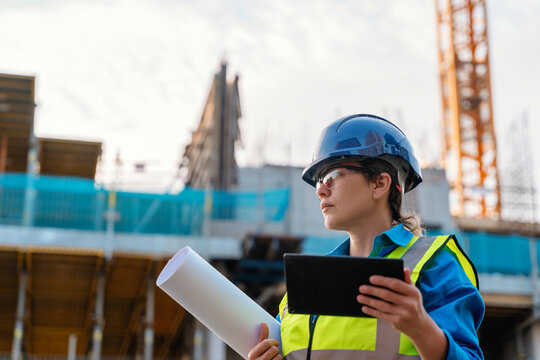 Portrait of Female construction supervisor reviews plans while ensuring safety protocols followed at busy building site with cranes and workers present, focused on efficient project management